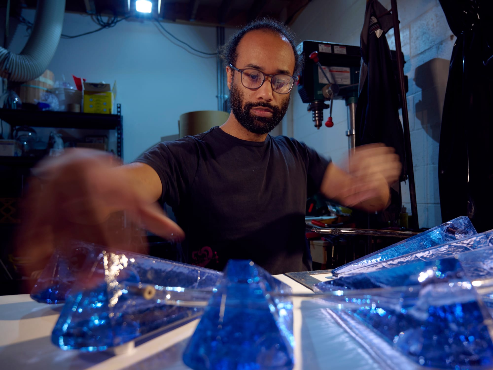 A man in glasses and a black shirt moves his hands while adjusting blue glass pieces