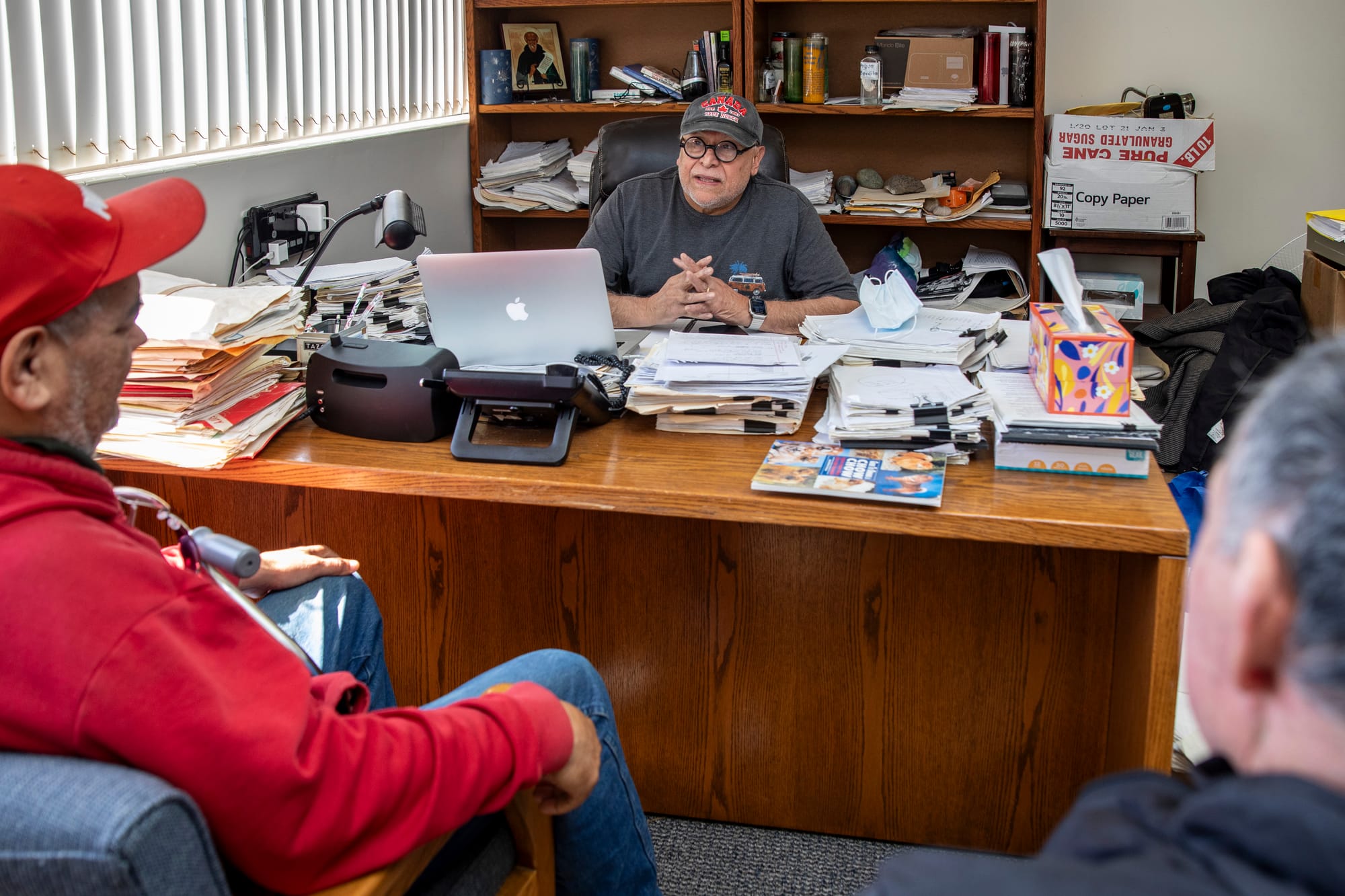 Three men sit in a room around a desk filled with papers.