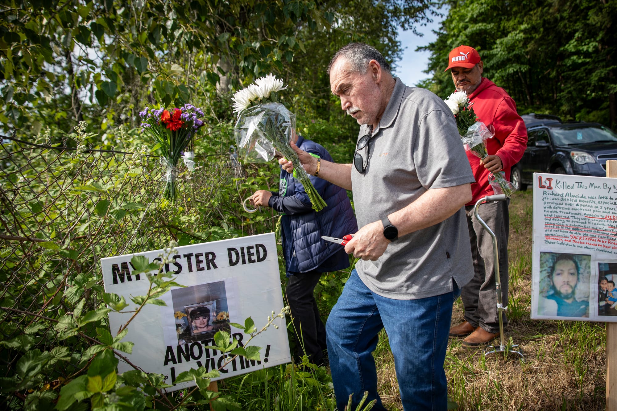 A man in a gray shirt holds flowers in one hand and scissors in the other. Two signs on stakes surround him.