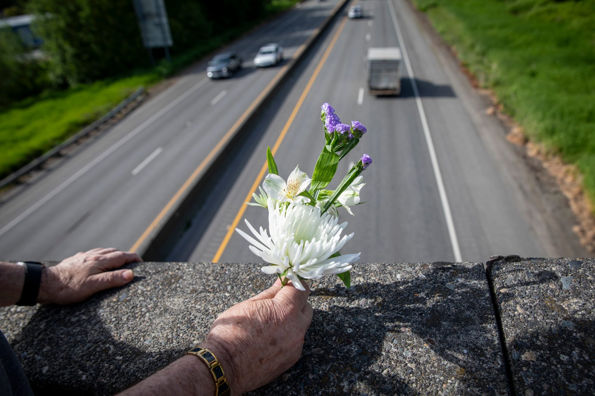 A hand holding purple and white flowers rests on a bridge over pass with cars underneath.