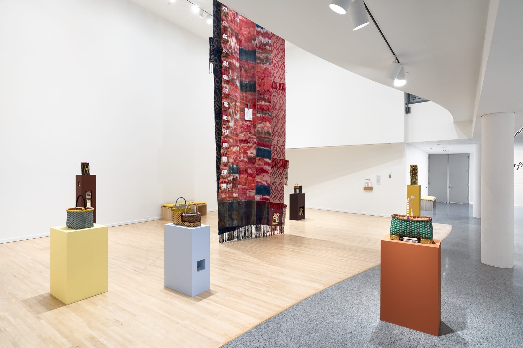 A white gallery with a large tapestry hanging at center, surrounded by small plinths with baskets on top