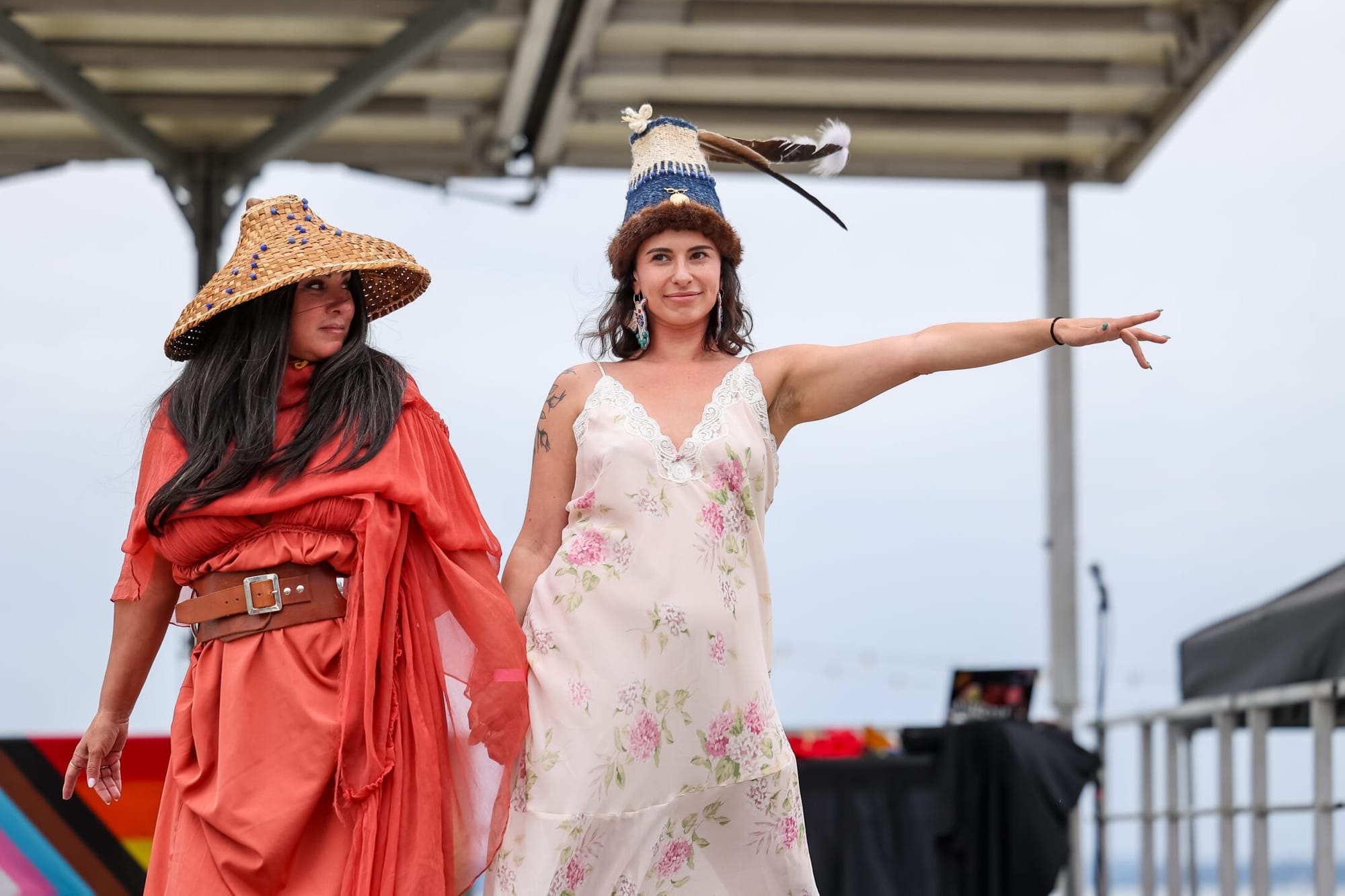 Two women strike a pose on an outdoor stage, both in dresses and woven hats