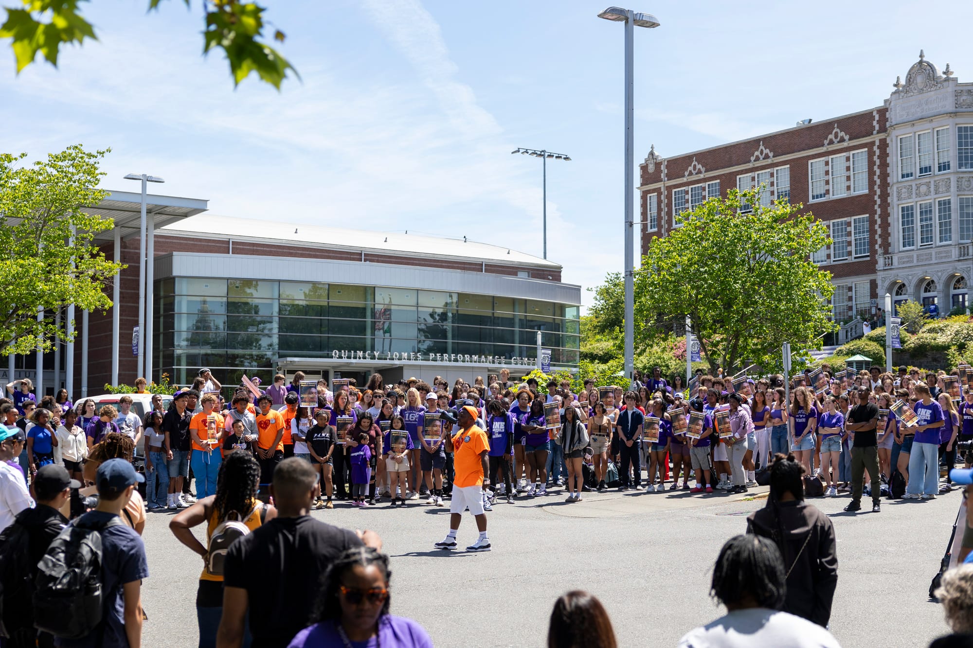 a man in an orange shirt speaks at a rally surrounded by hundreds of students and community members