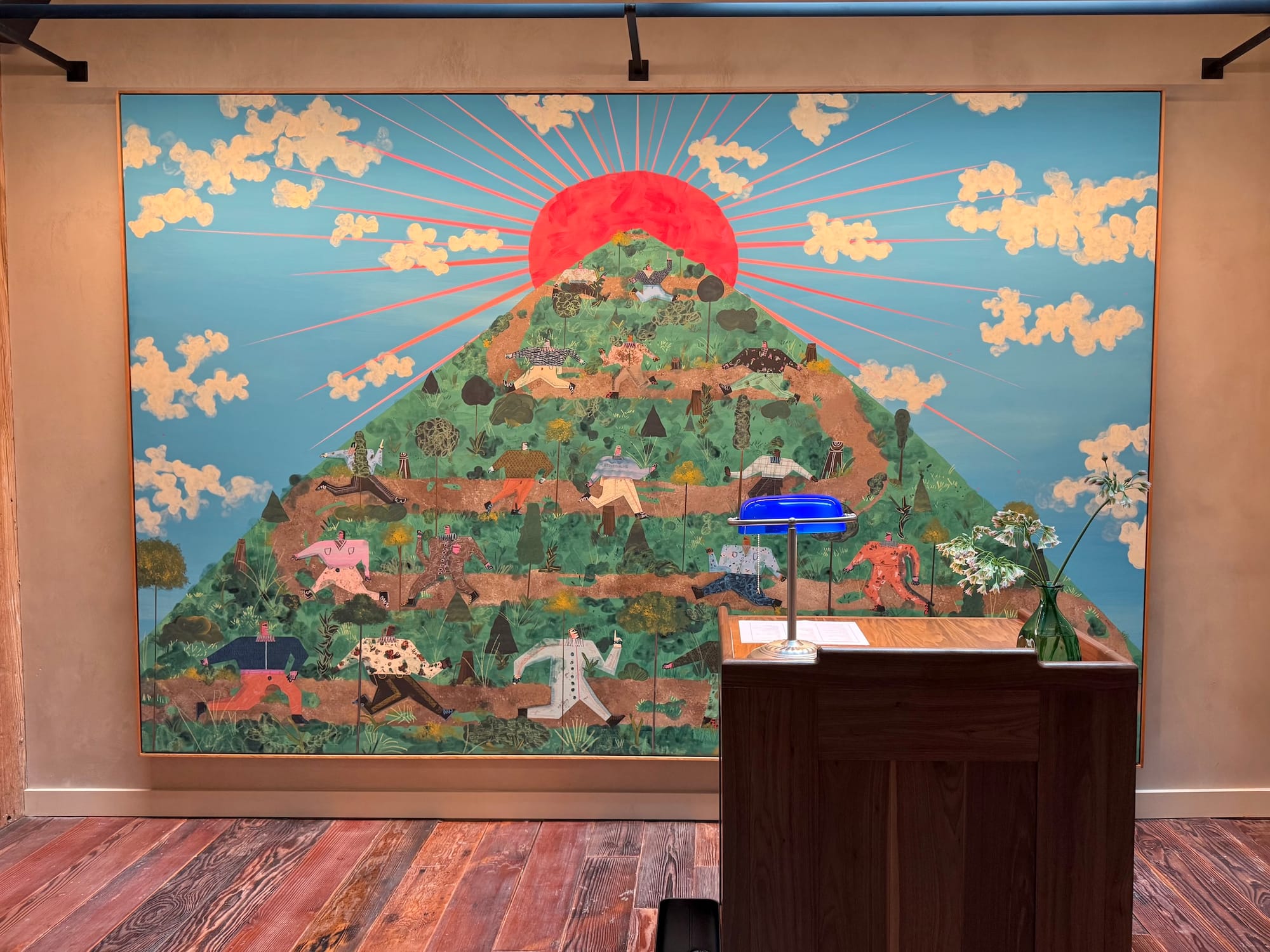 Photo of a large painting hung behind a small desk with a blue banker's lamp on it. Painting is of a green mountain with red sun rising behind