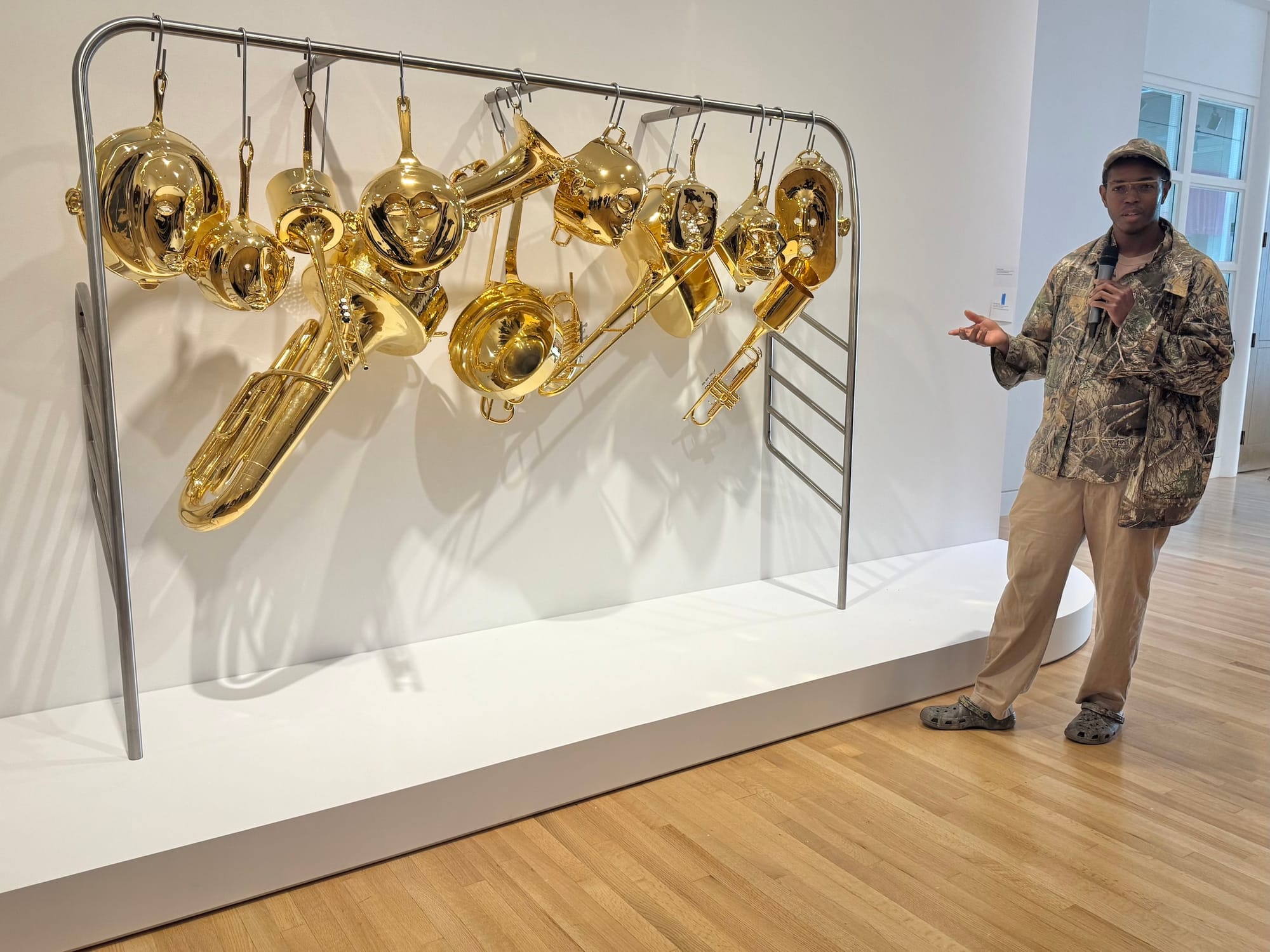A man with a mic gestures toward a sculpture featuring unusually shaped, bright gold horn instruments hanging from a subway rail.