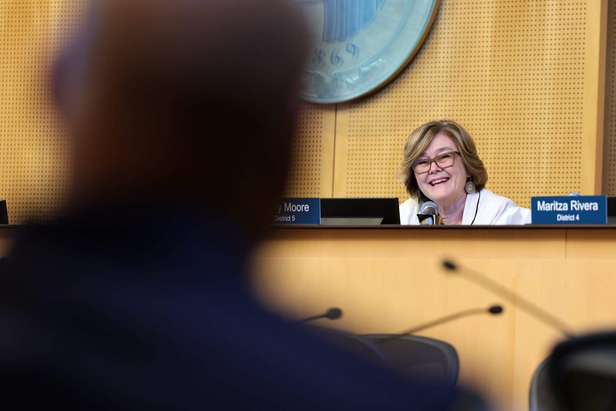 a white woman sits in council chambers behind the dais. she is smiling