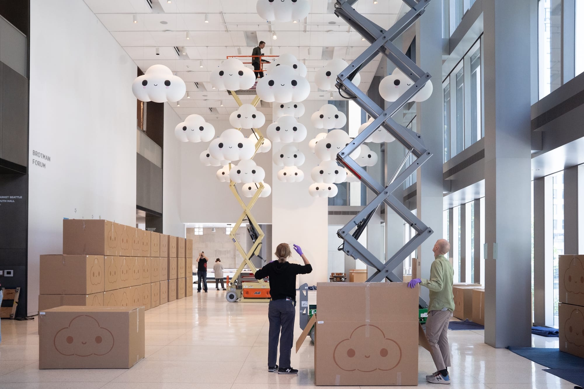 a tall windowed gallery with many cardboard boxes stacked on one side; workers look up as they hang smiling clouds from the ceiling