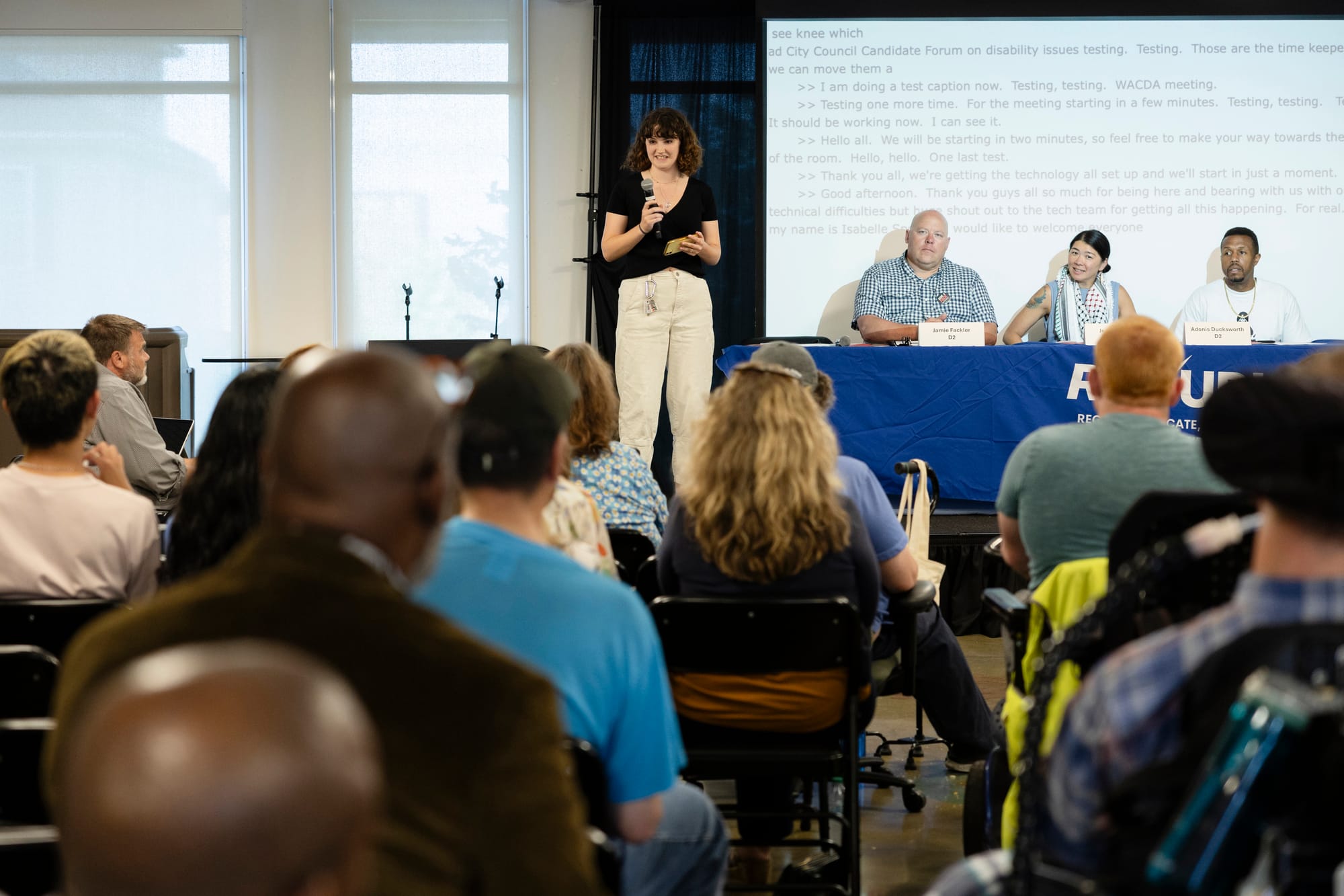 a white woman stands with a mic. a white man, asian woman and black man sit at a table next to her. in the foreground an audience sits in chairs.