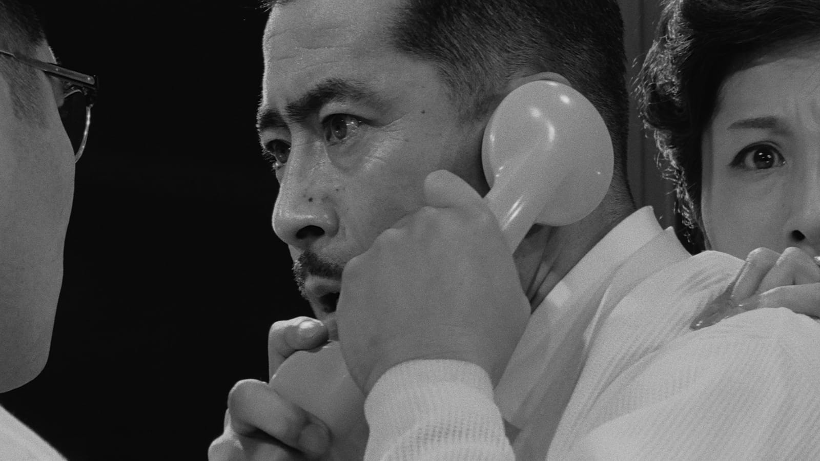 Black and white film still in which a worried man is clutching a telephone to his ear