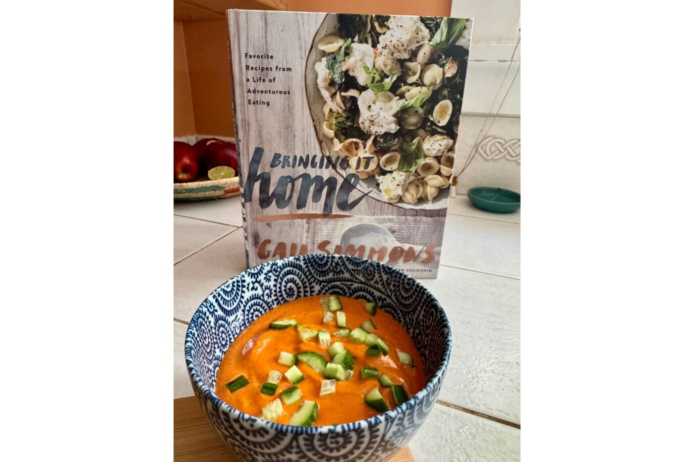 A bowl of Gazpacho soup sits in front of a standing cookbook. Text on the cookbook reads "Favorite Recipes from a life of adventurous eating; 'Bringing it Home'; Gail Simmons"