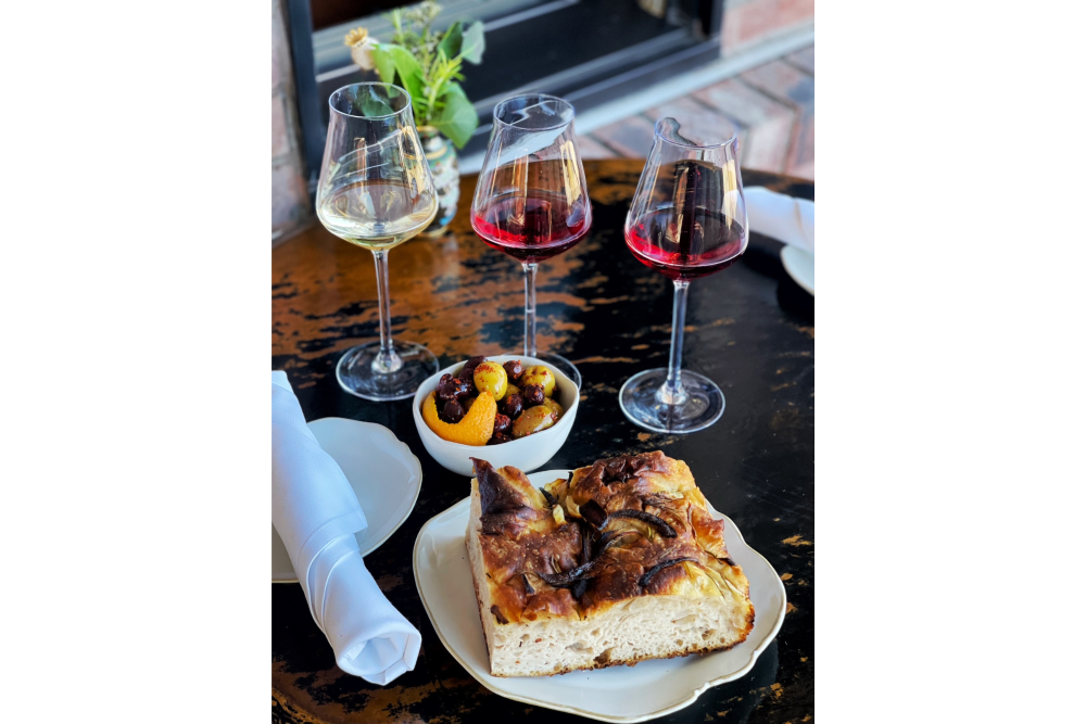 A few glasses of wine, a bowl of mixed fruit and a piece of focaccia bread on a plate.