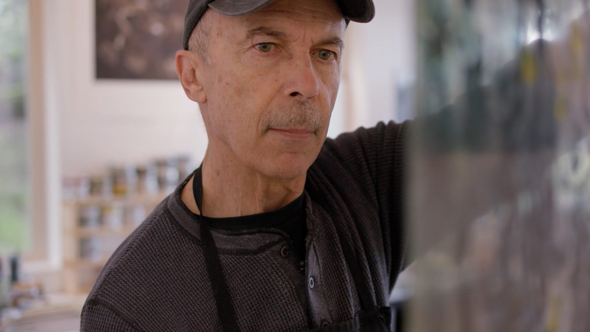 A man wearing a cap and printer's apron holds up a painted glass and looks at it.