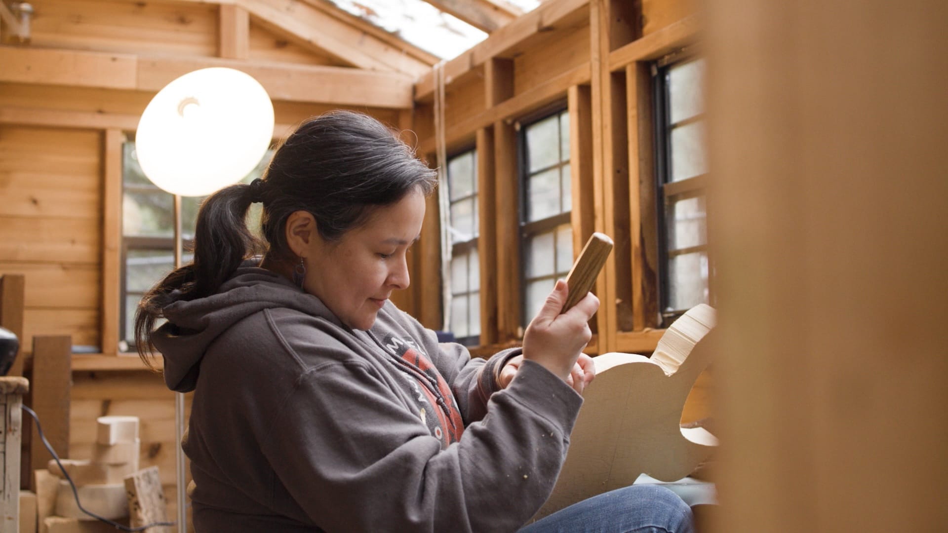 A ponytailed woman in a wood walled studio uses a chisel to carve a flat piece of wood.
