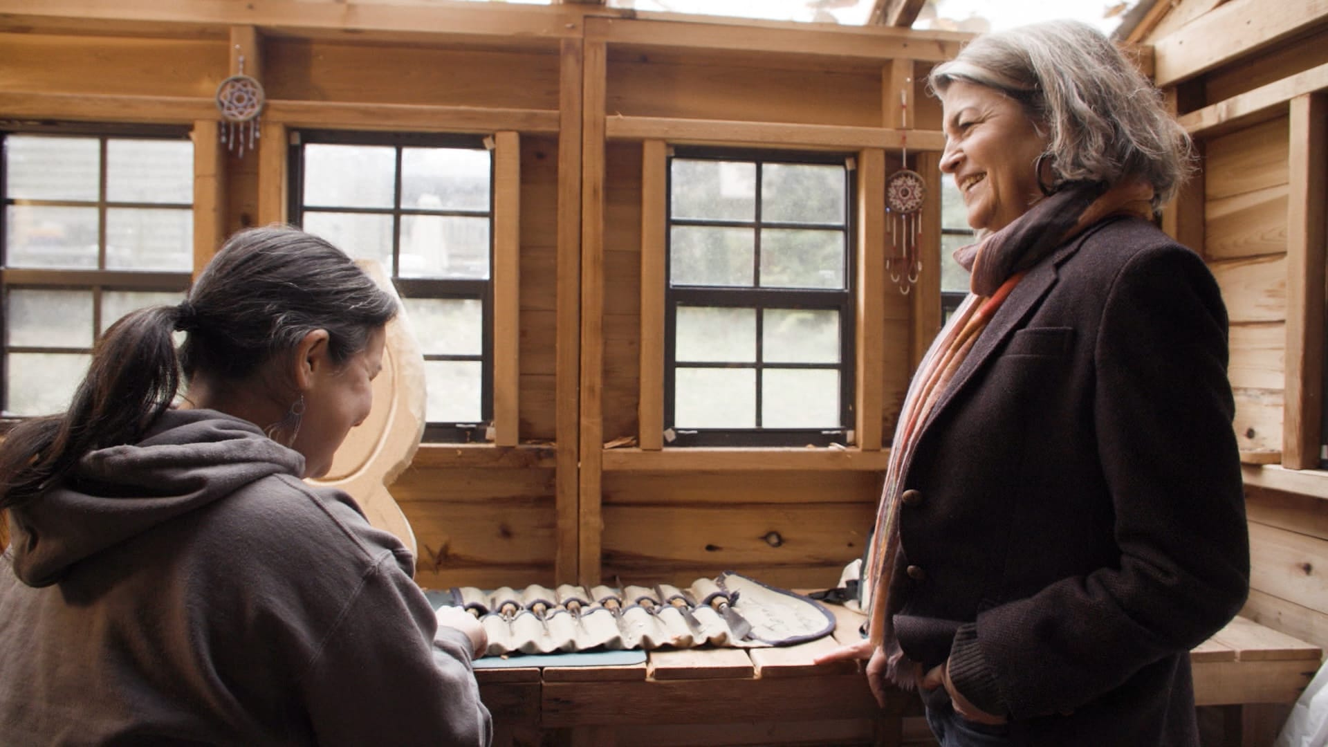 Two women face each other smiling in front of windows and a counter with satchel full of carving tools.
