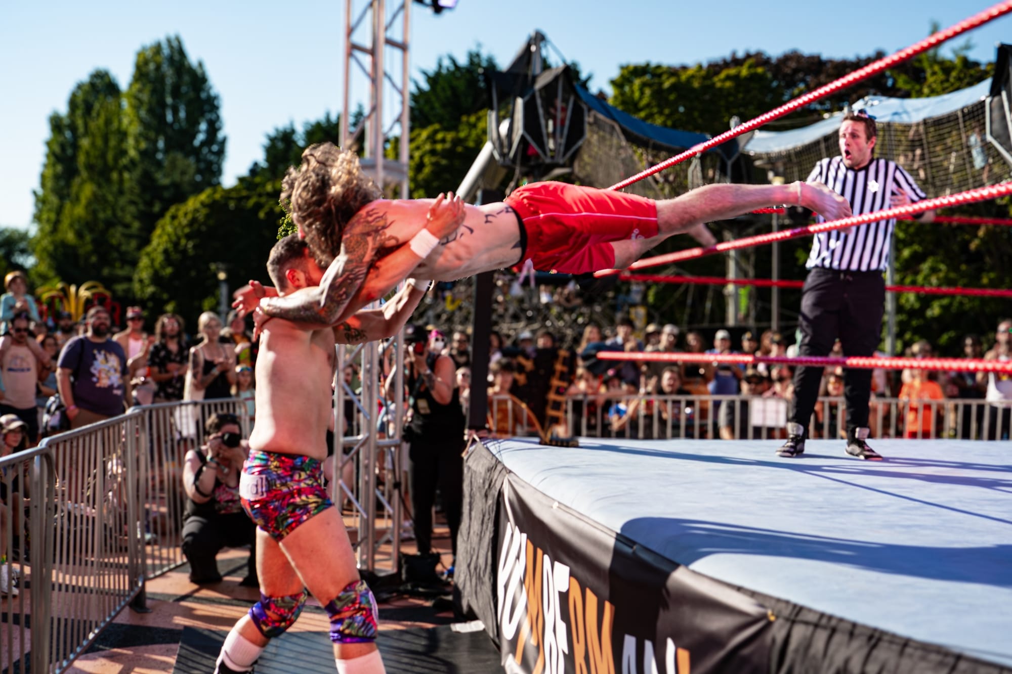 Photo of an outdoor pro-wrestling match where one man in shorts is leaping on another.