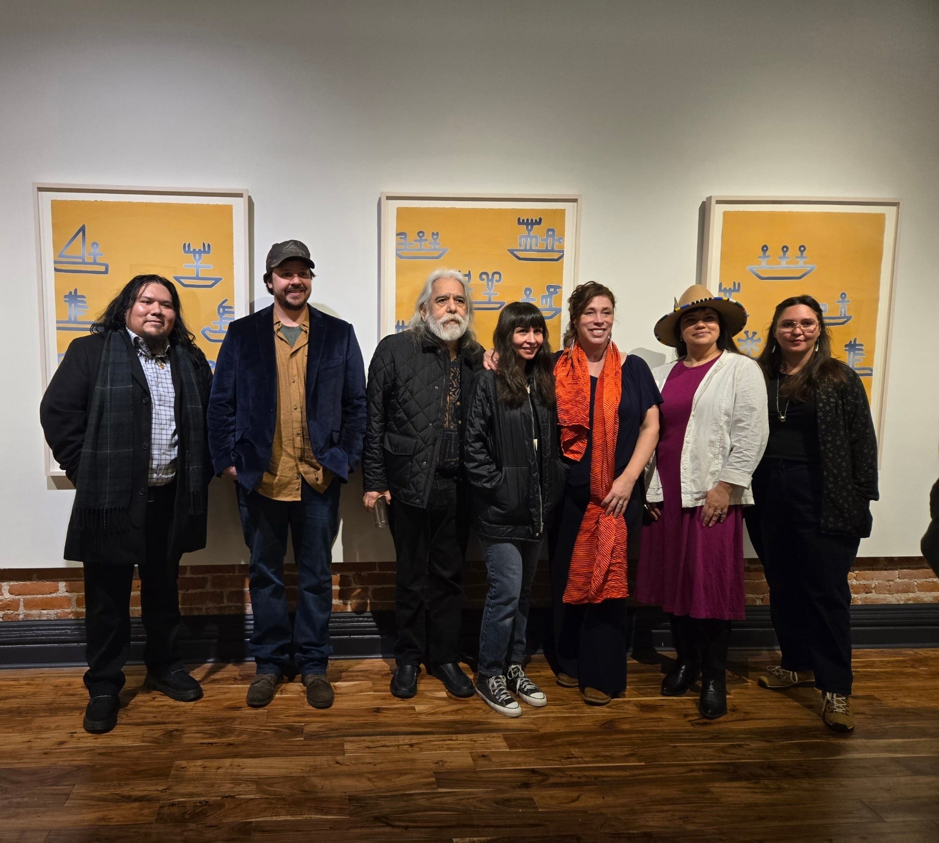 A photo of seven artists posing in front of art at a gallery.