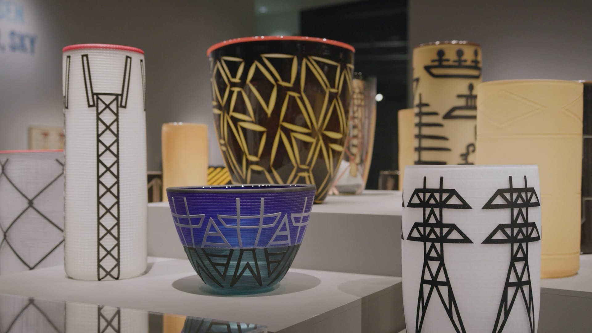 Photo of a gallery display of glass vessels adorned with graphic symbols suggesting high voltage towers, canoes and trees.
