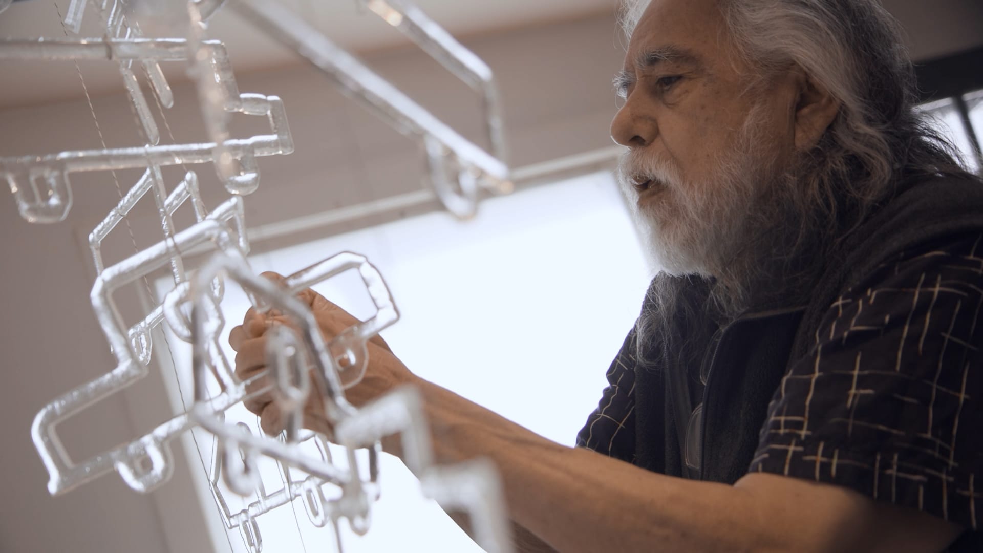 A man with long white hair and beard hangs clear glass iconography on thin wires in a home