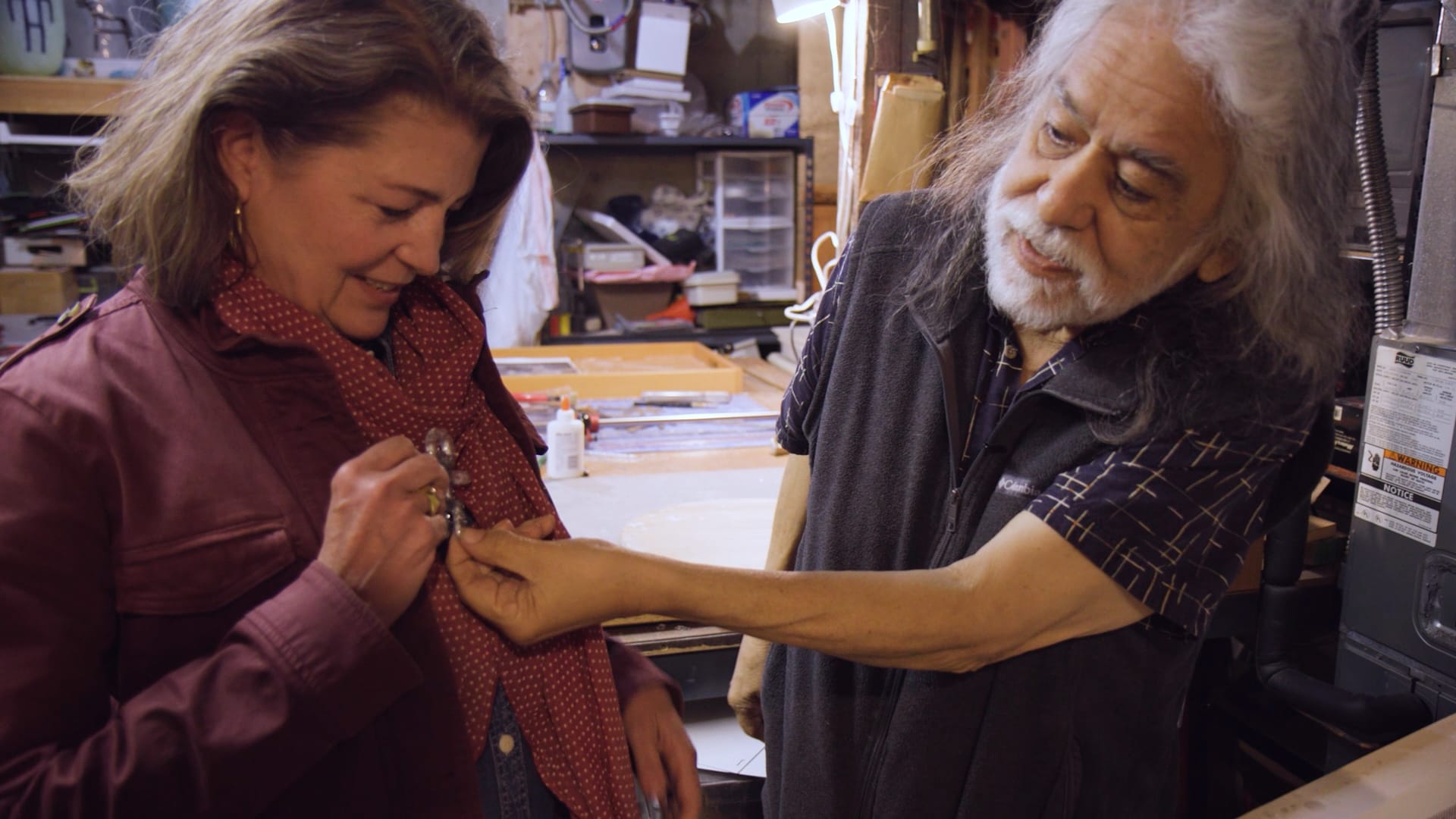 A man presents a woman with a small glass pendant in a room crowded with art supplies.