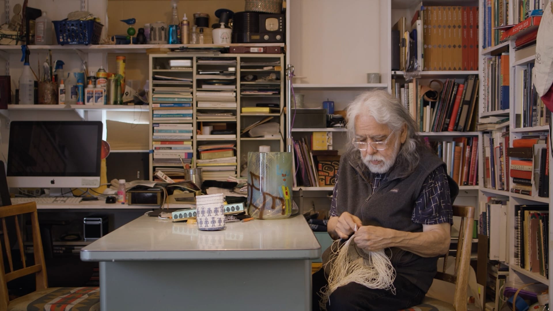 A man with long gray hair and glasses weaves a basket in a room crowded with shelves of art books