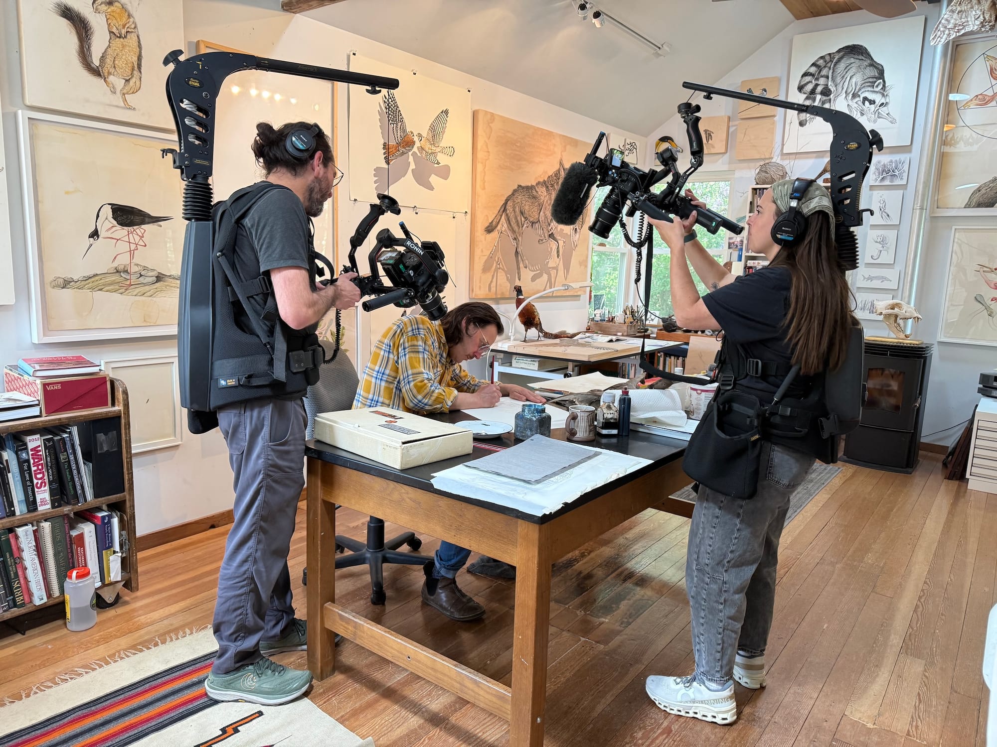 Photo of an artist's studio, with an artist at a drawing table and two camera people filming from above.