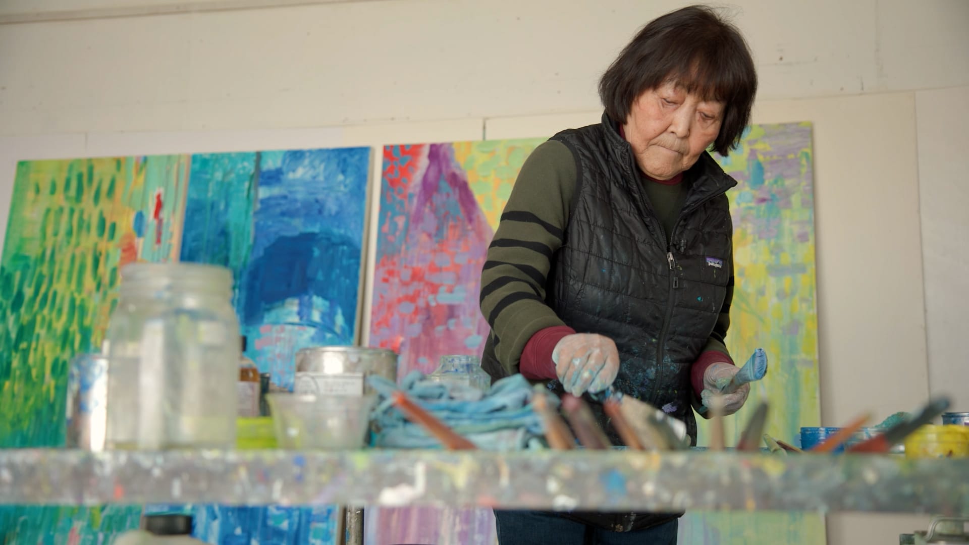 A woman with short dark hair mixes paint in an artist studio. Four tall abstract paintings lean on the wall behind