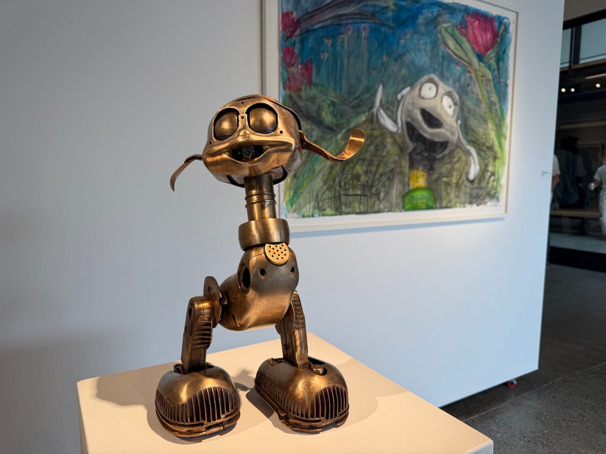 A strange bronze creature-like toy on a pedestal in front of a painting.