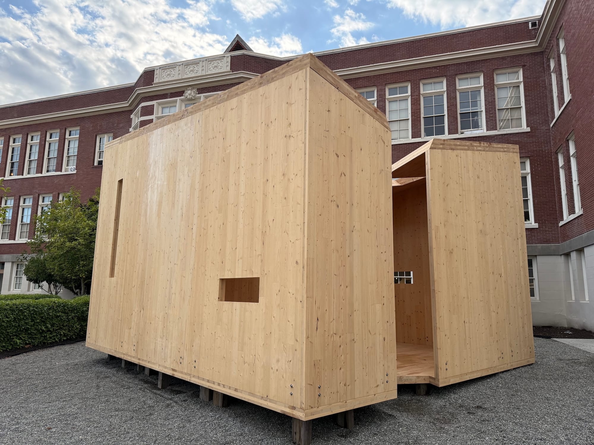 A modern angular wooden structure sits in front of a classical brick building.
