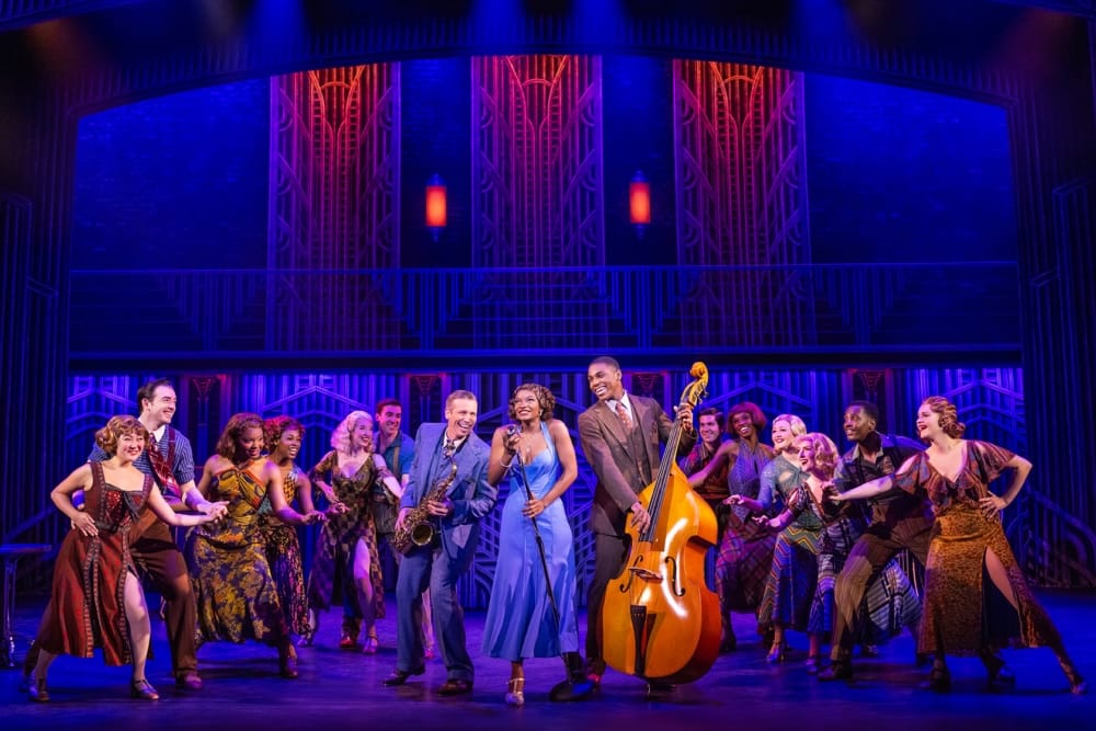 A chorus of actors on a purple-tinted stage with a singer, saxophonist and bass player at front