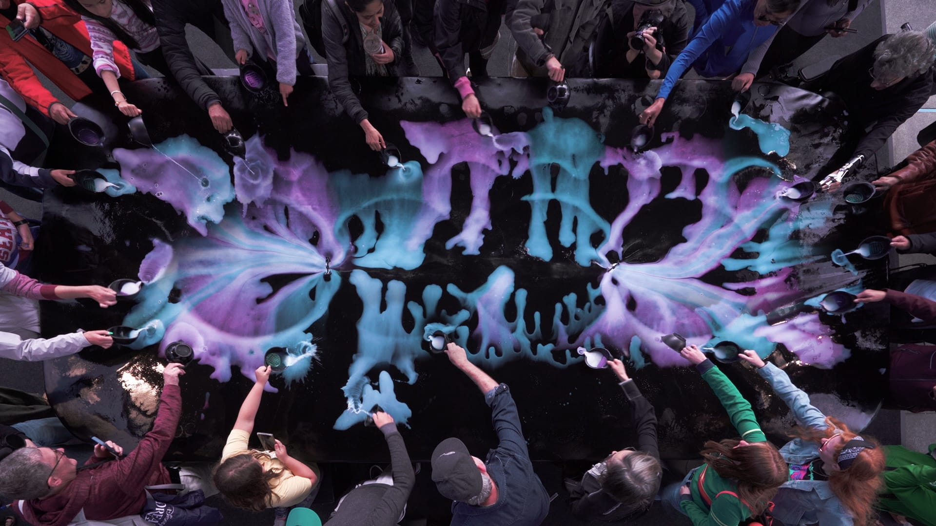 In a birds-eye photo, many people surround a large canvas and pour vibrant paint on it