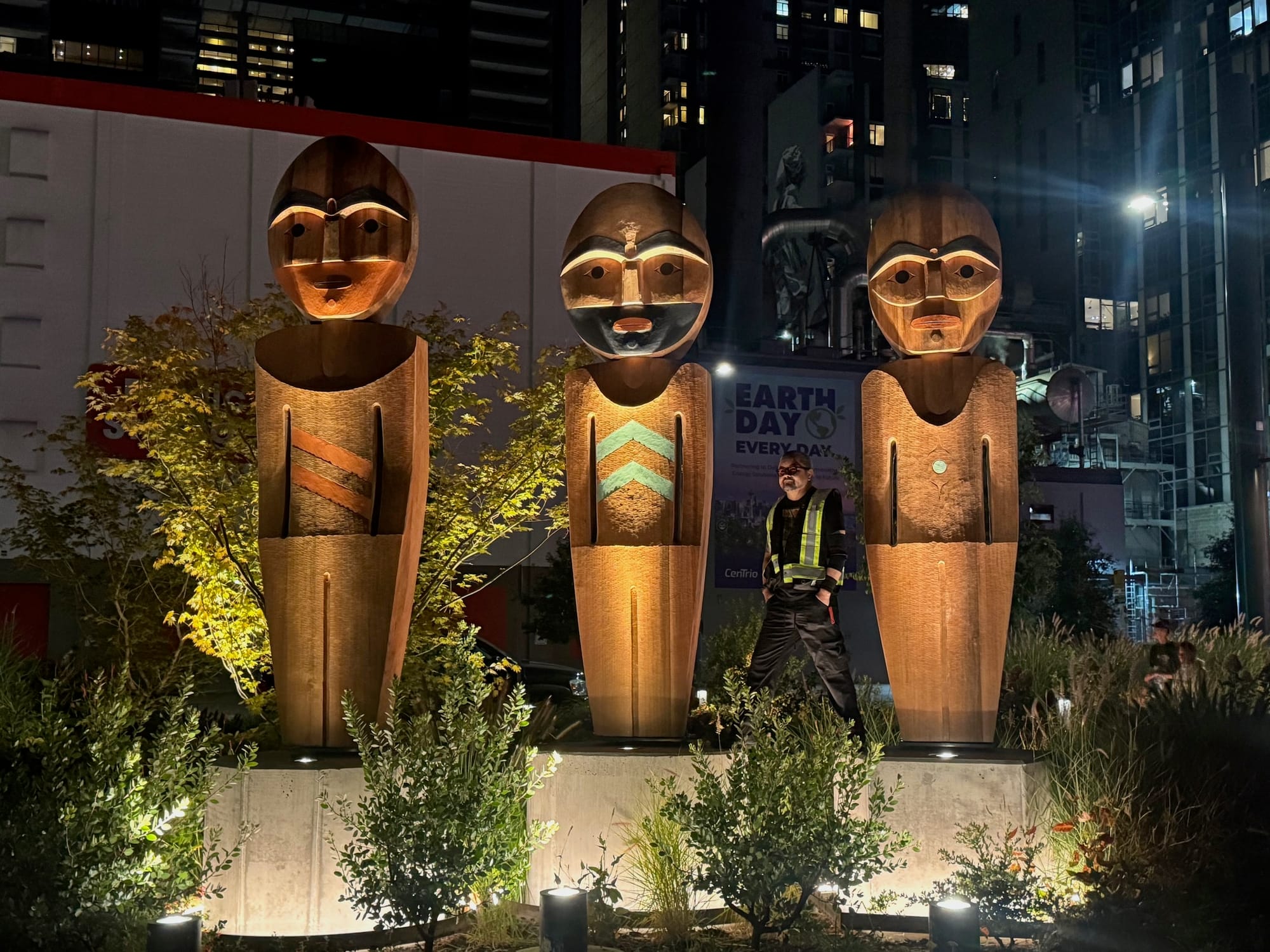 Three wooden figures carved in Coast Salish style, stand on concrete pedestals lit at night.
