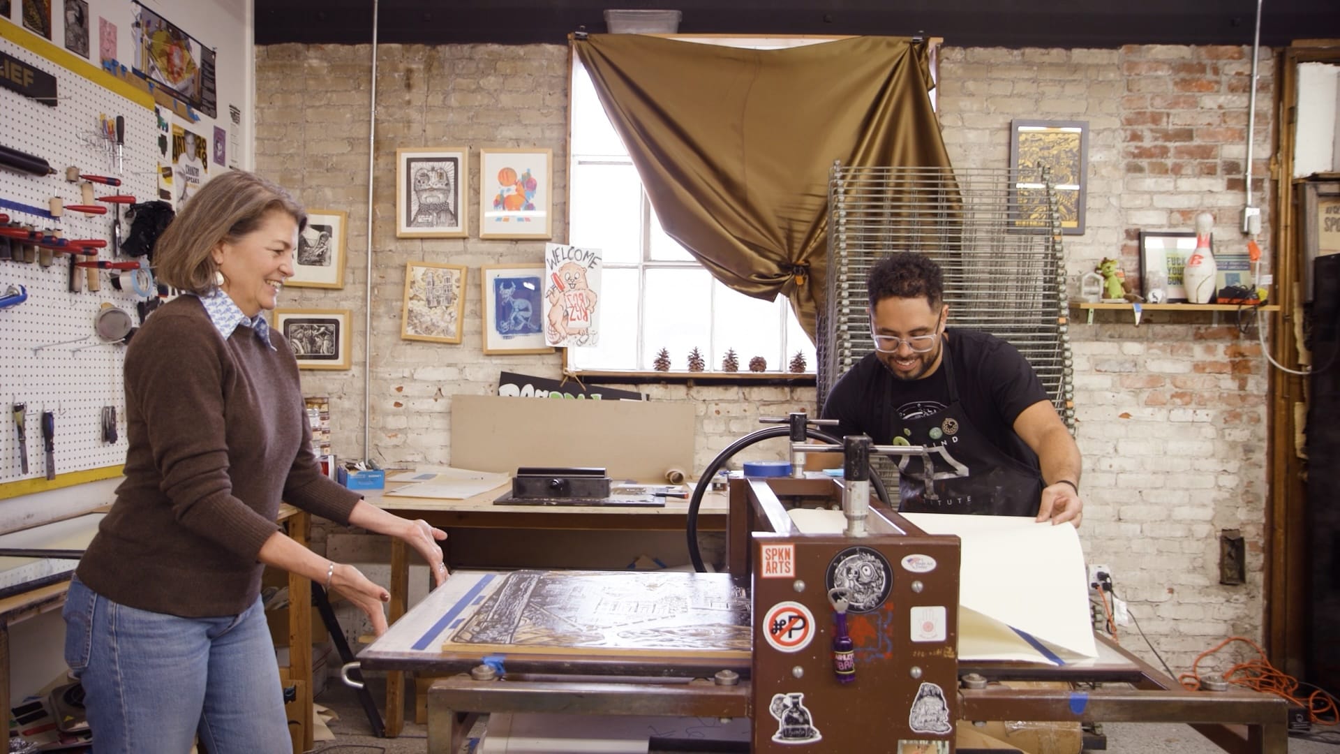 A man and a woman work to pull a print on an industrial printing press in a studio.