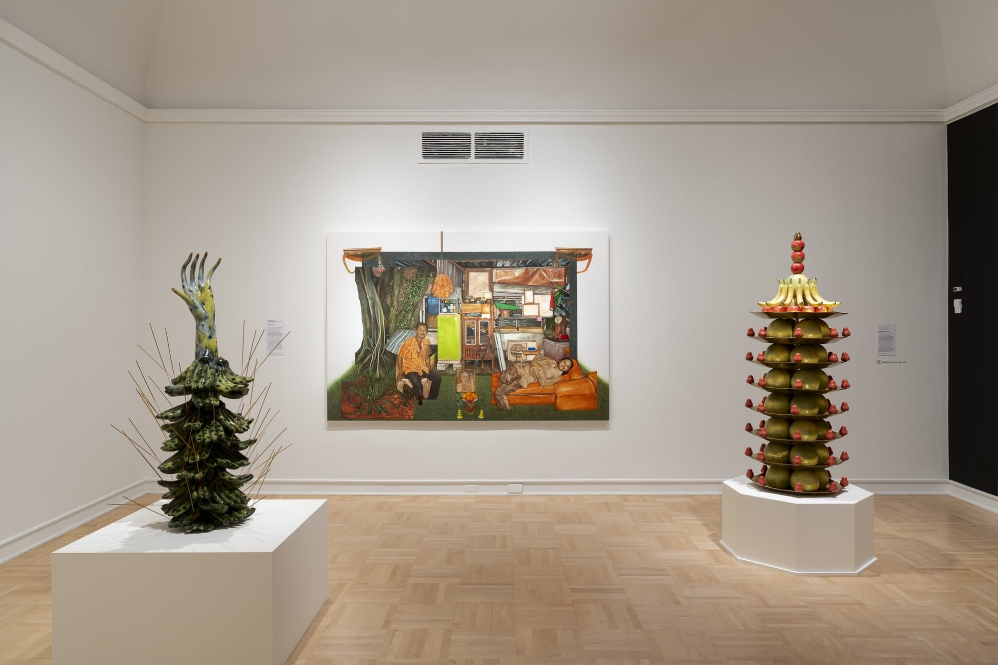A white-walled gallery featuring two ceramic altars filled with fake fruit surrounding a painting of an interior room.