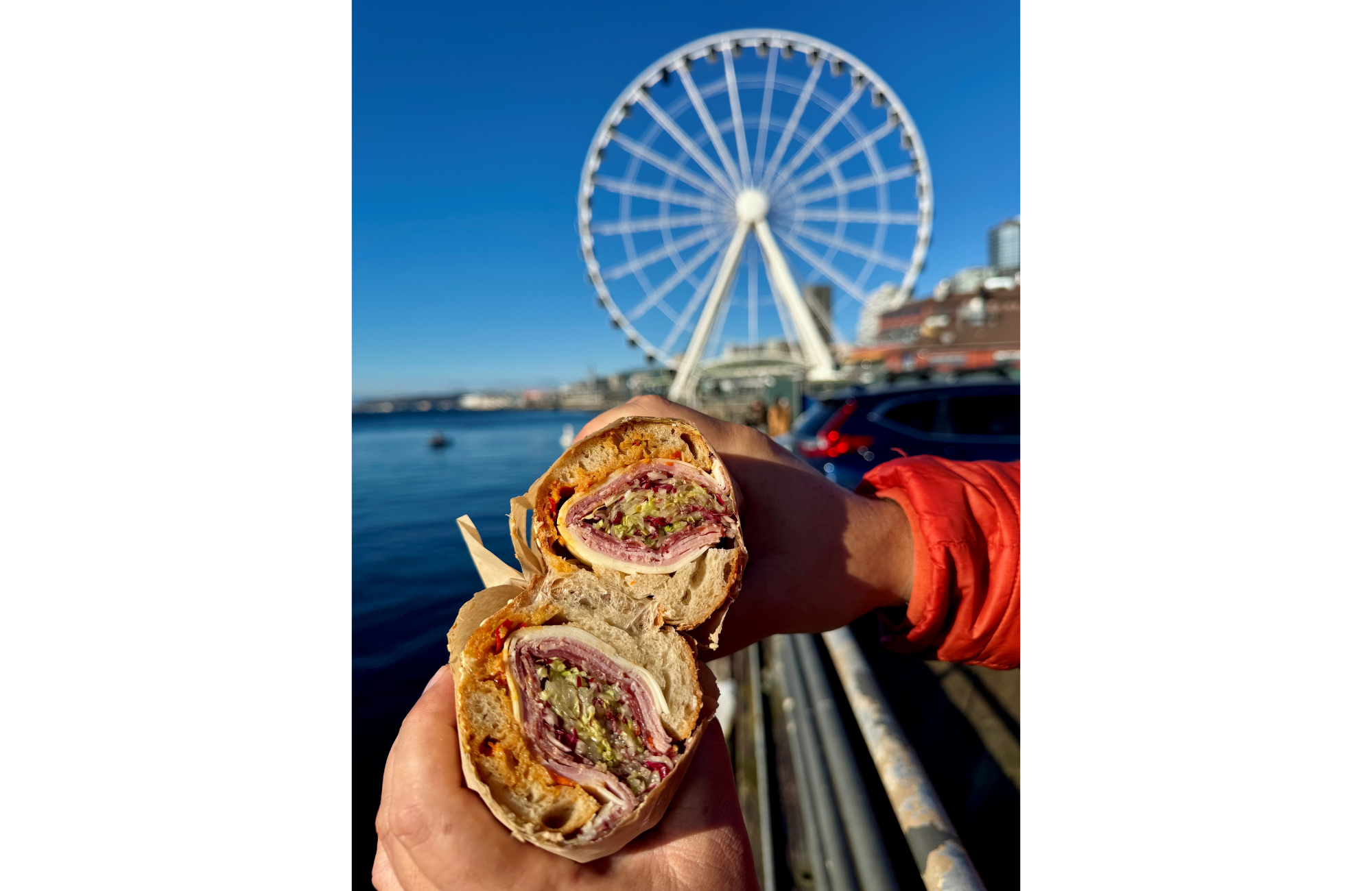 Two people hold up Italian sandwiches in front of a view of the waterfront and a ferris wheel.