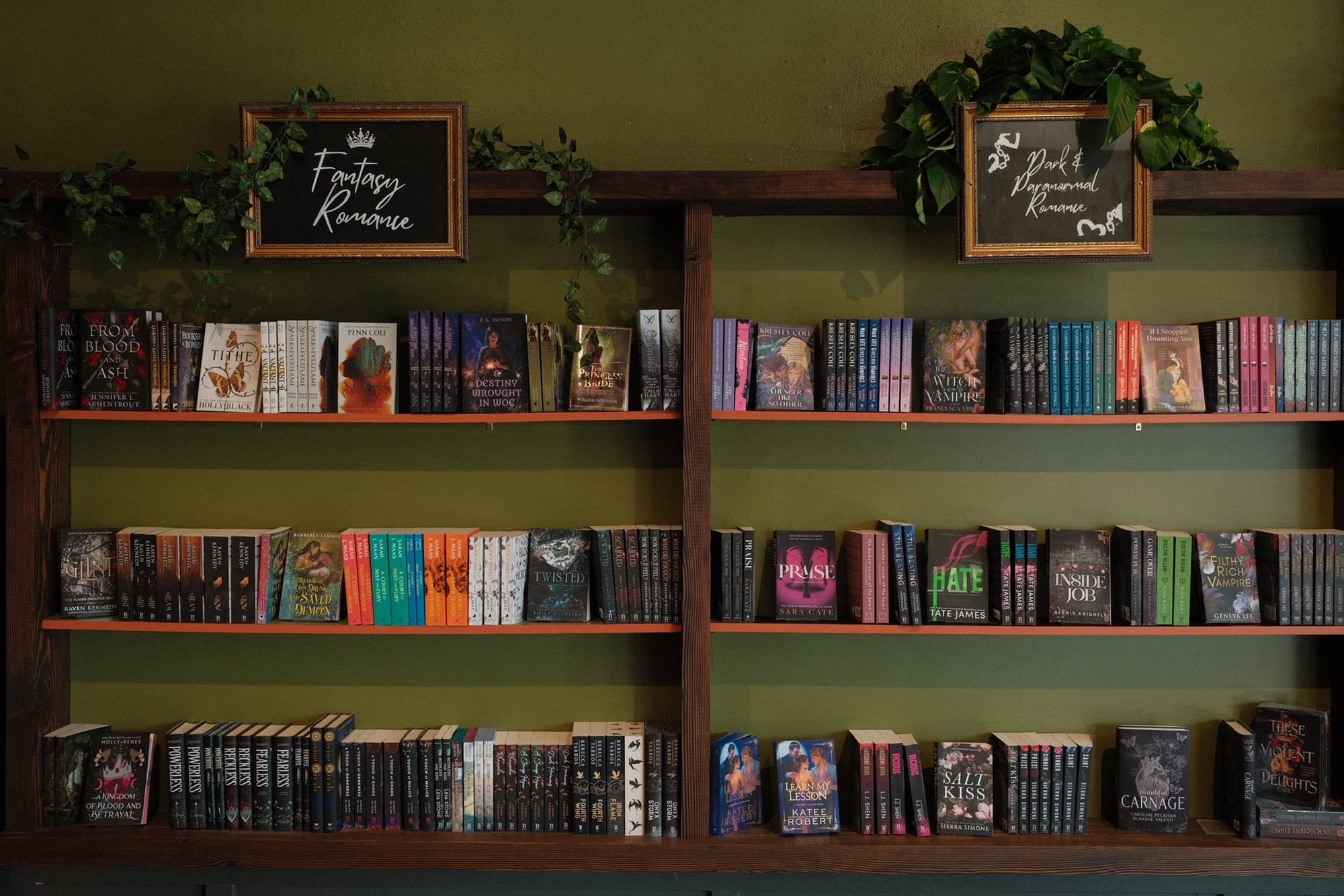 Photo of al tall wooden bookshelf against a dark olive wall, stacked with many romance books.