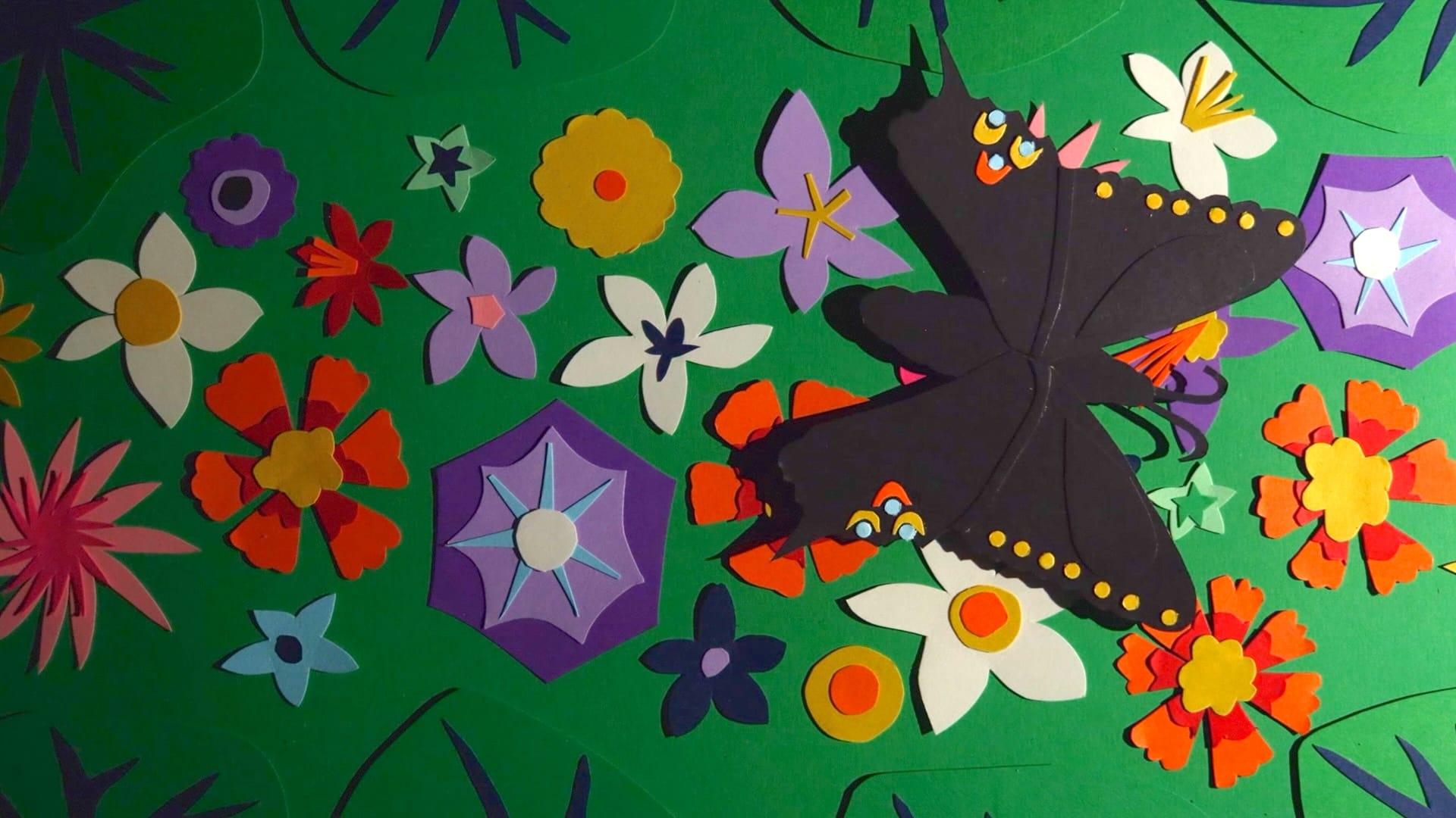 A still from an animated movie featuring cut-paper flowers and a black paper butterfly on a green background.