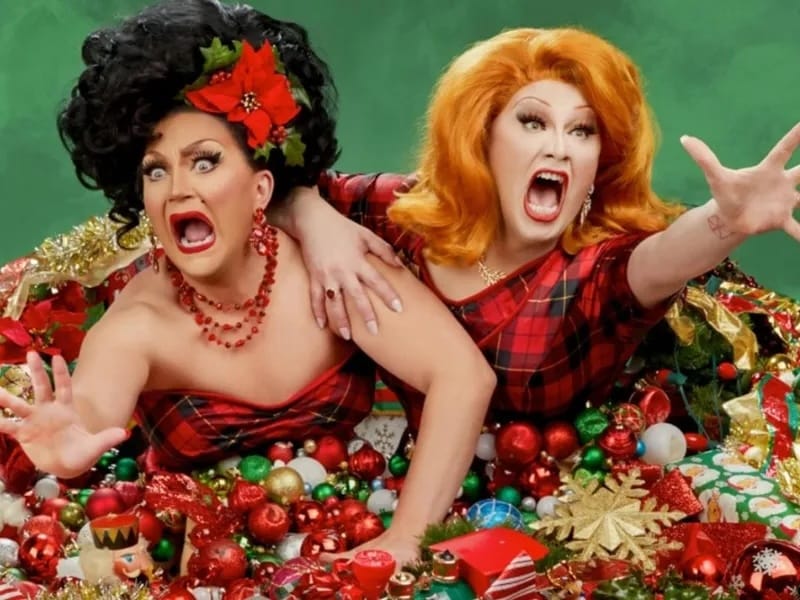 Two drag queens crawl out from a pile of holiday ornaments.