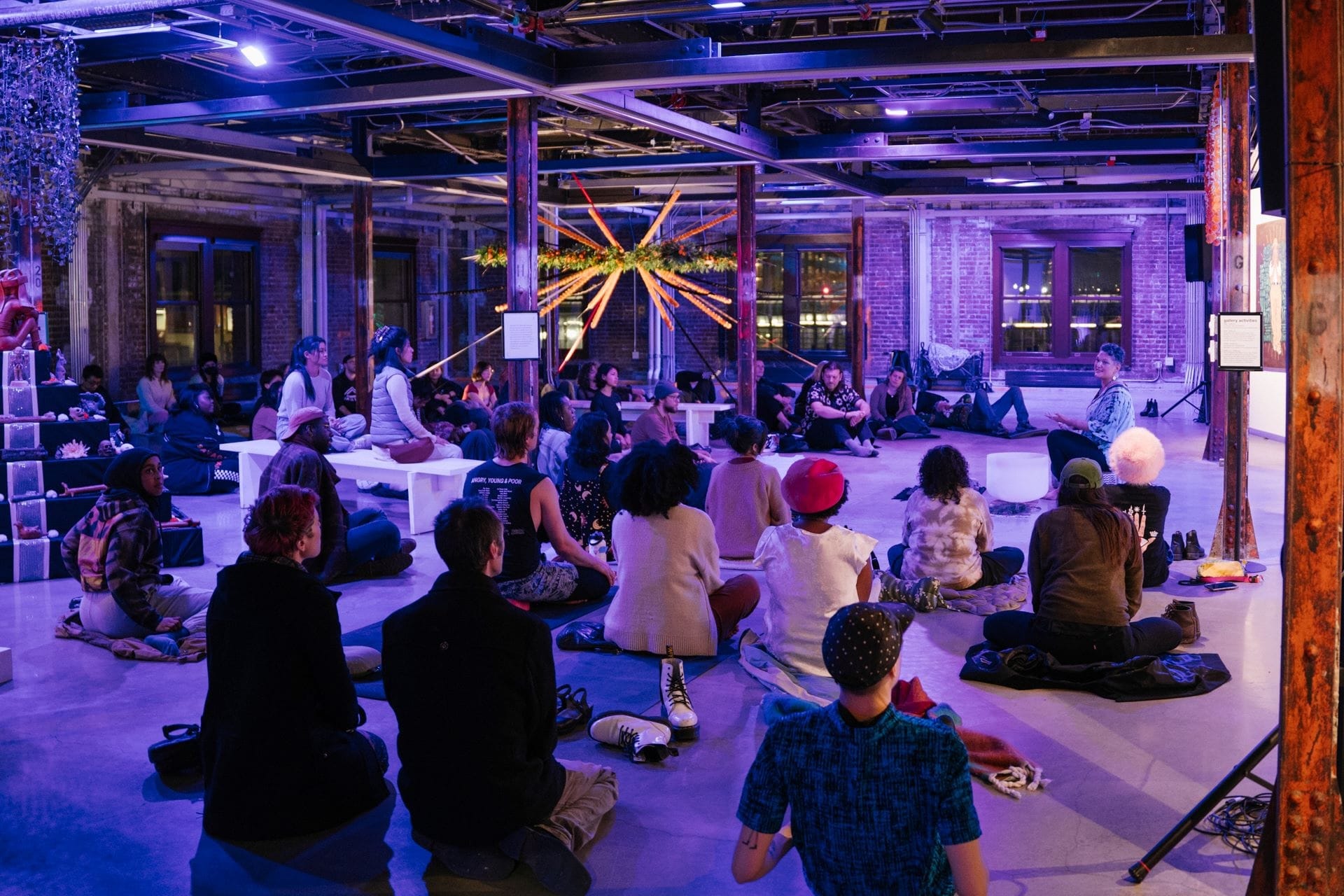 In a brick-walled warehouse space, a group of people sit on the floor around a large lit star.