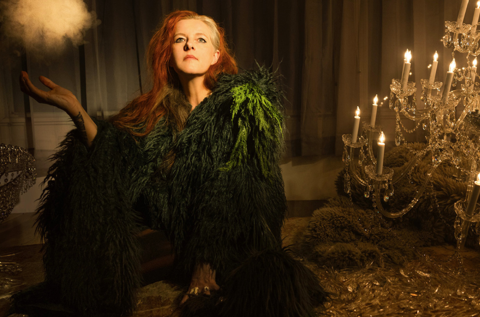 Musician Neko Case wears a mossy robe and is crouched on a floor in a dimly lit room next to a fallen chandelier