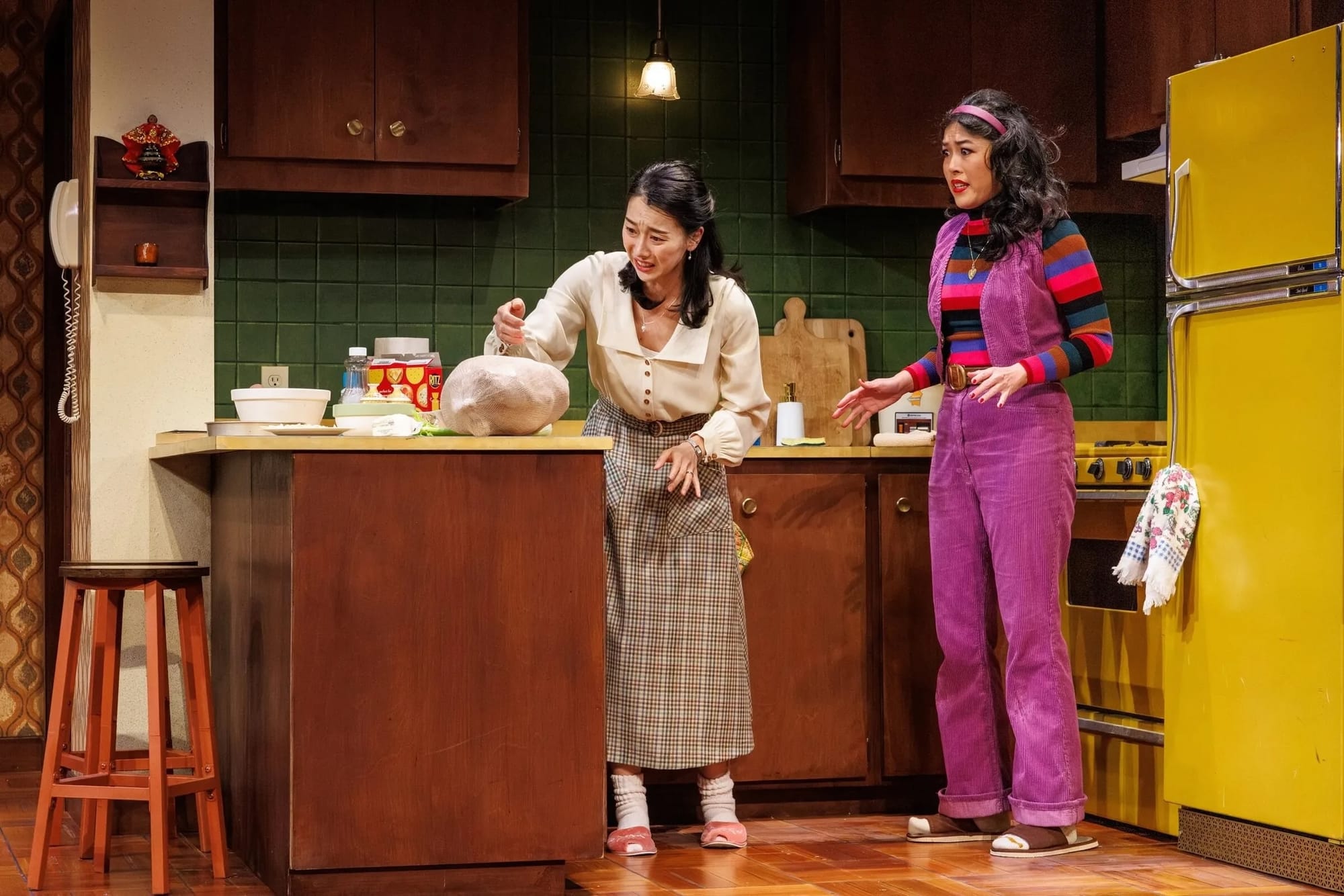 A stage play with two women in a kitchen wearing 1970s clothes and looking alarmed by a frozen chicken