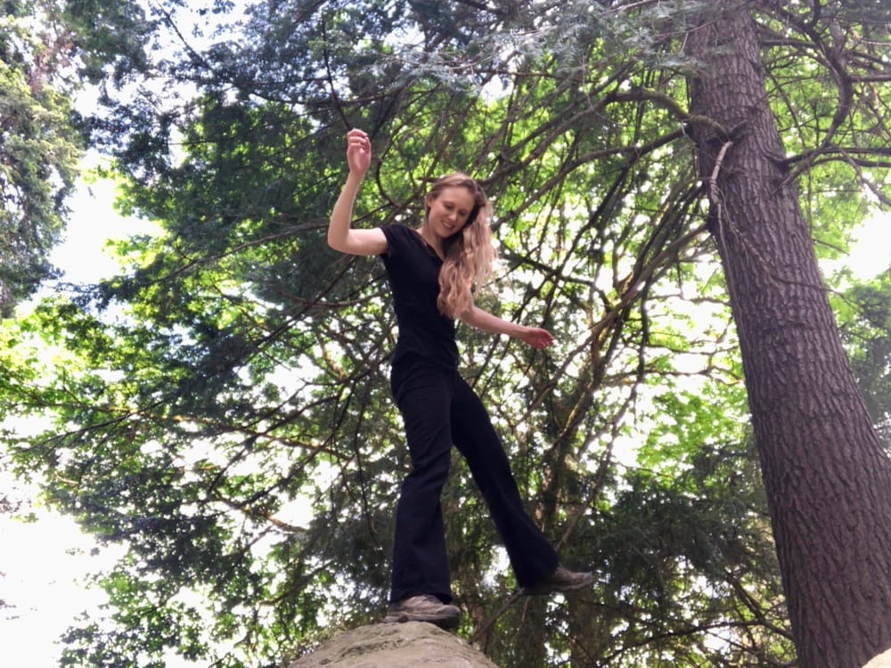 A woman in black pants and black shirt balances on a large rock in the woods. 