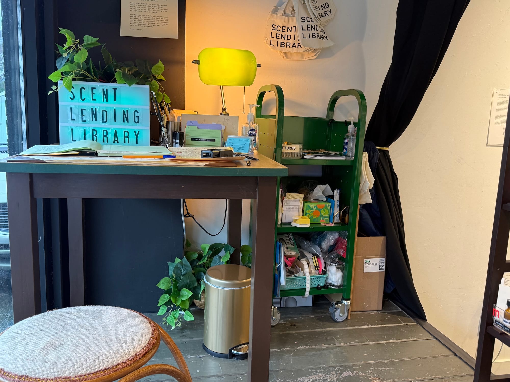 Photo of a small desk with a sign reading Scent Lending Library next to a green shelving unit