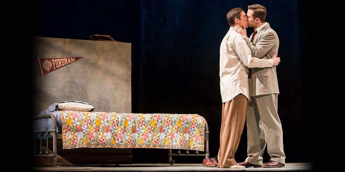 On a theatrically lit stage, two men in suits stand and kiss in front of a single bed. 