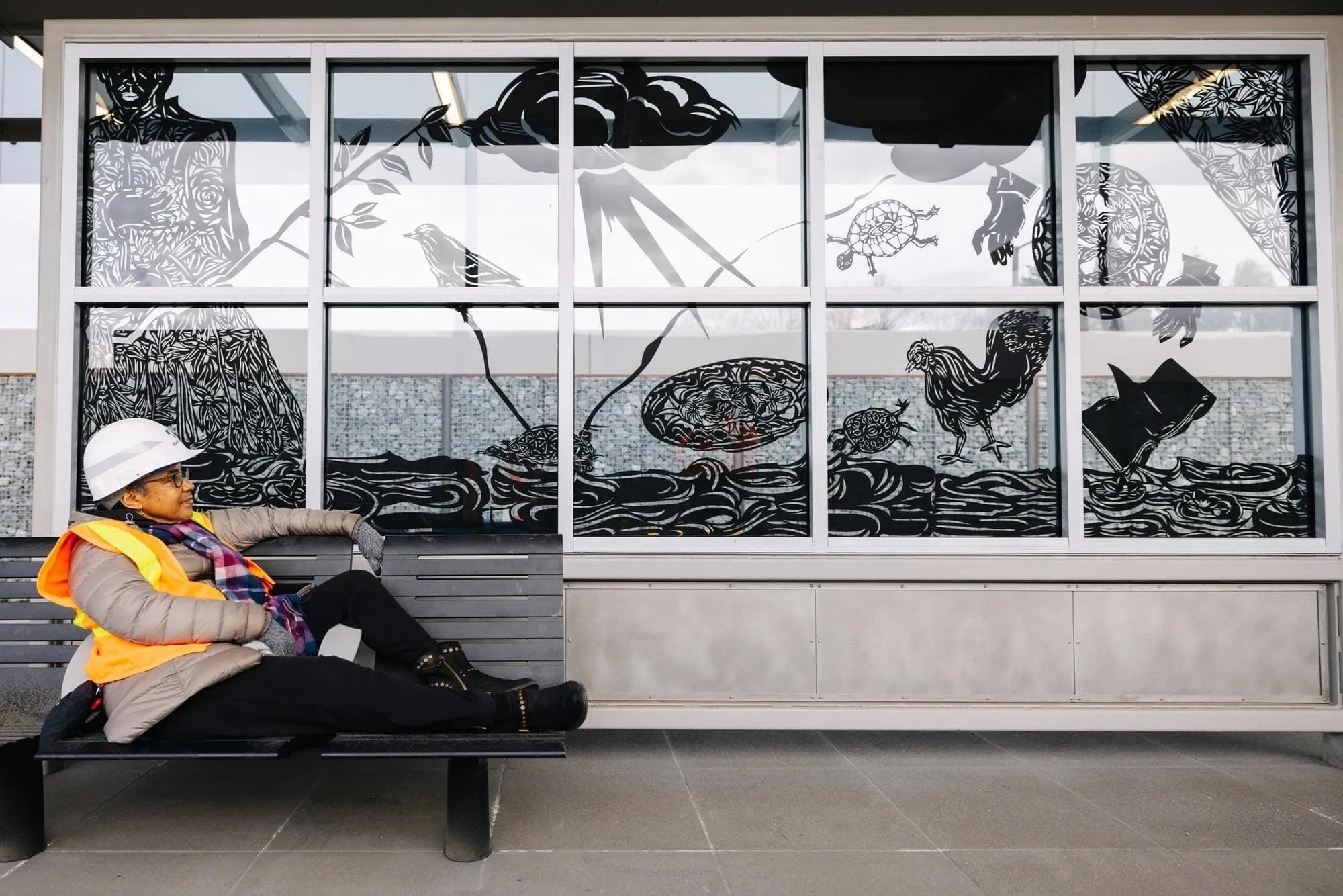 A woman in a hard hat sits on a bench in front of a segmented glass panel with panes decorated with nature scenes