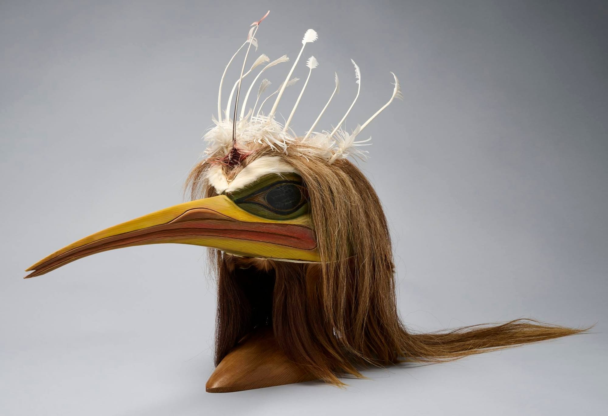 A gallery image of a Coast Salish mosquito mask, with a long, thin beak, long horse hair and stripped feathers poking out of its crown
