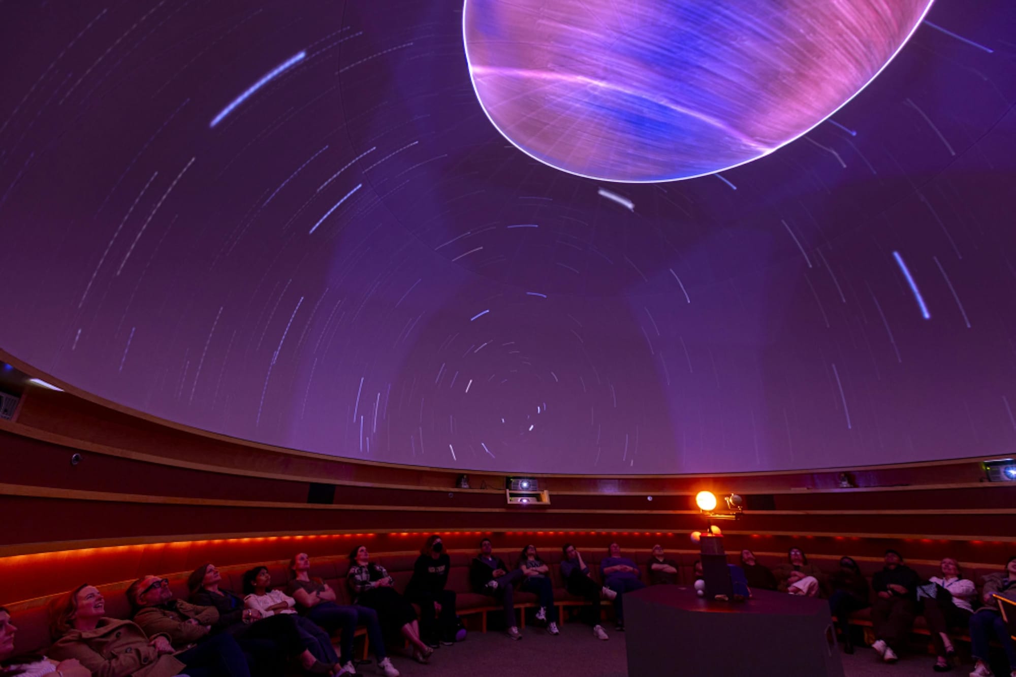 A photo of the inside of a planetarium, with amphitheatre seating and a starscape above seated visitors