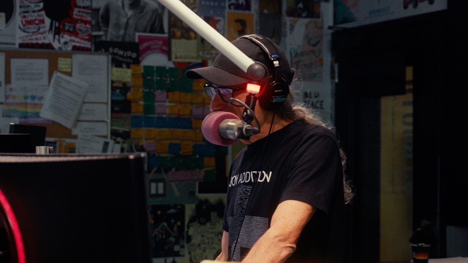 A DJ wearing a baseball cap, glasses and headphones talks from behind a microphone.