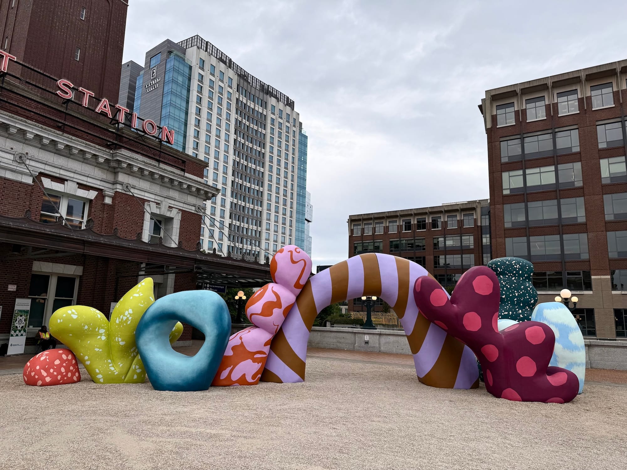 Photo of a large, colorful, abstract sculpture on a plaza outside a train station.
