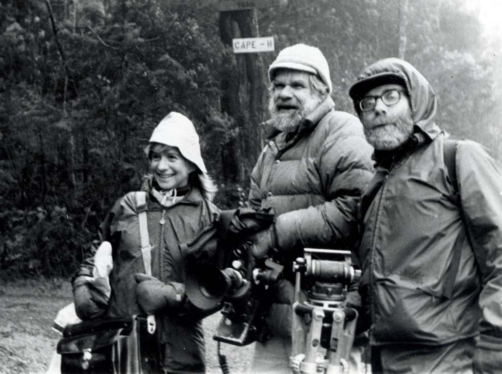 A vintage black-and-white photo of a woman and two men holding heavy camera gear out in the woods