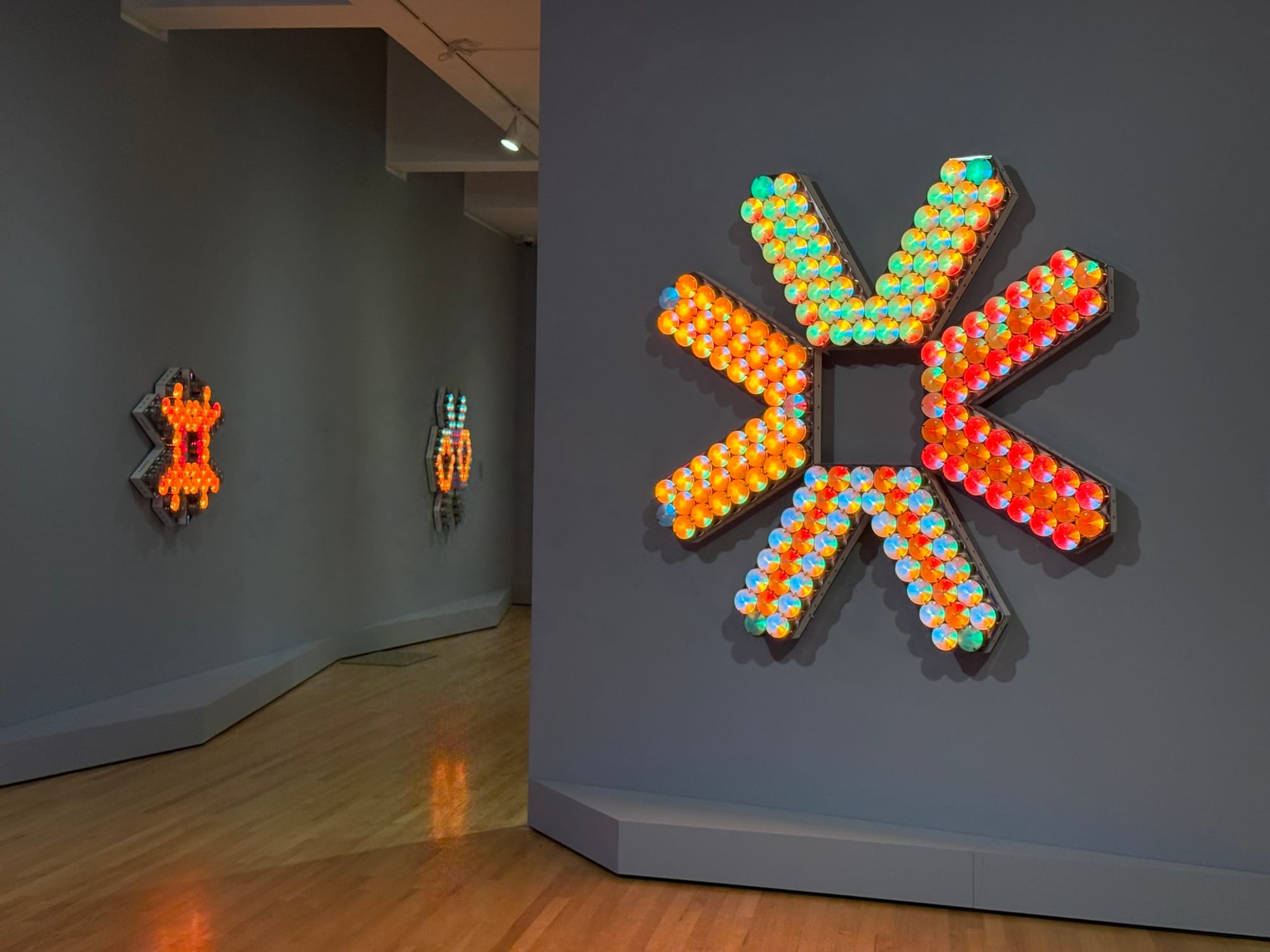 Exhibit featuring three geometric light installations on gray walls.